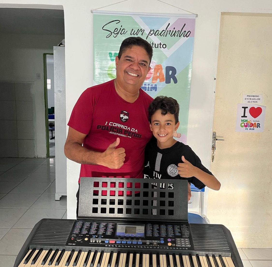 Um menino e um educador sorrindo atrás de um teclado, ao lado do cartaz do apadrinhamento
